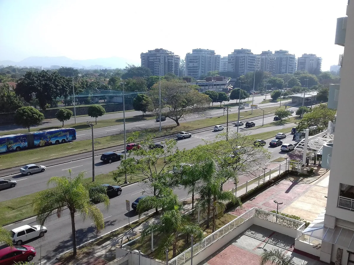 Avenida das Américas, na Barra da Tijuca, com pistas largas, canteiro arborizado e prédios residenciais ao fundo.
