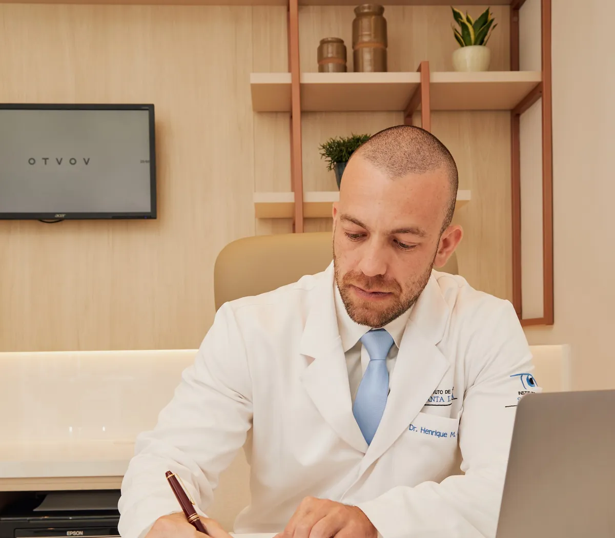 Dr. Henrique Leber, de jaleco, sentado à mesa do consultório, escrevendo com uma caneta ao lado de um notebook.