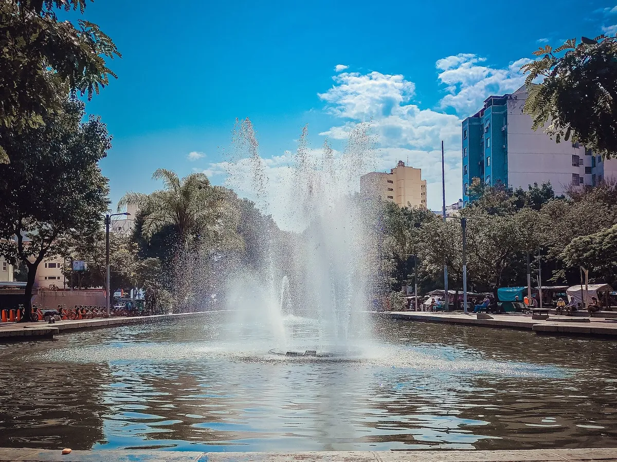 Chafariz com jato de água em um espelho d'água na Praça Saens Peña, com árvores e prédios ao fundo sob céu azul.
