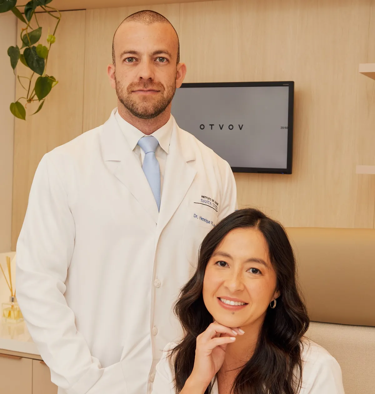Dr. Henrique Leber em pé e Dra. Nina Konichi sentada à frente, de jaleco, sorrindo no consultório.