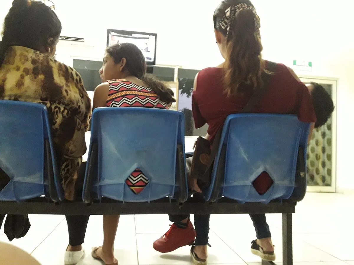 Três pessoas sentadas, de costas, em bancos azuis de uma sala de espera, olhando para um monitor.