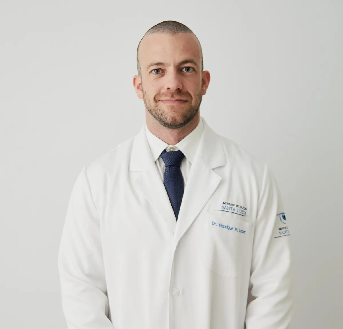 Dr. Henrique Leber, de jaleco branco e gravata azul-marinho, em pé diante de um fundo claro.