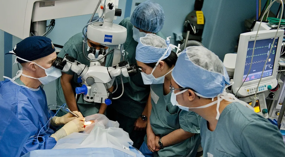 Equipe cirúrgica de máscara e touca ao redor de um microscópio cirúrgico durante uma cirurgia de catarata em sala de operação.