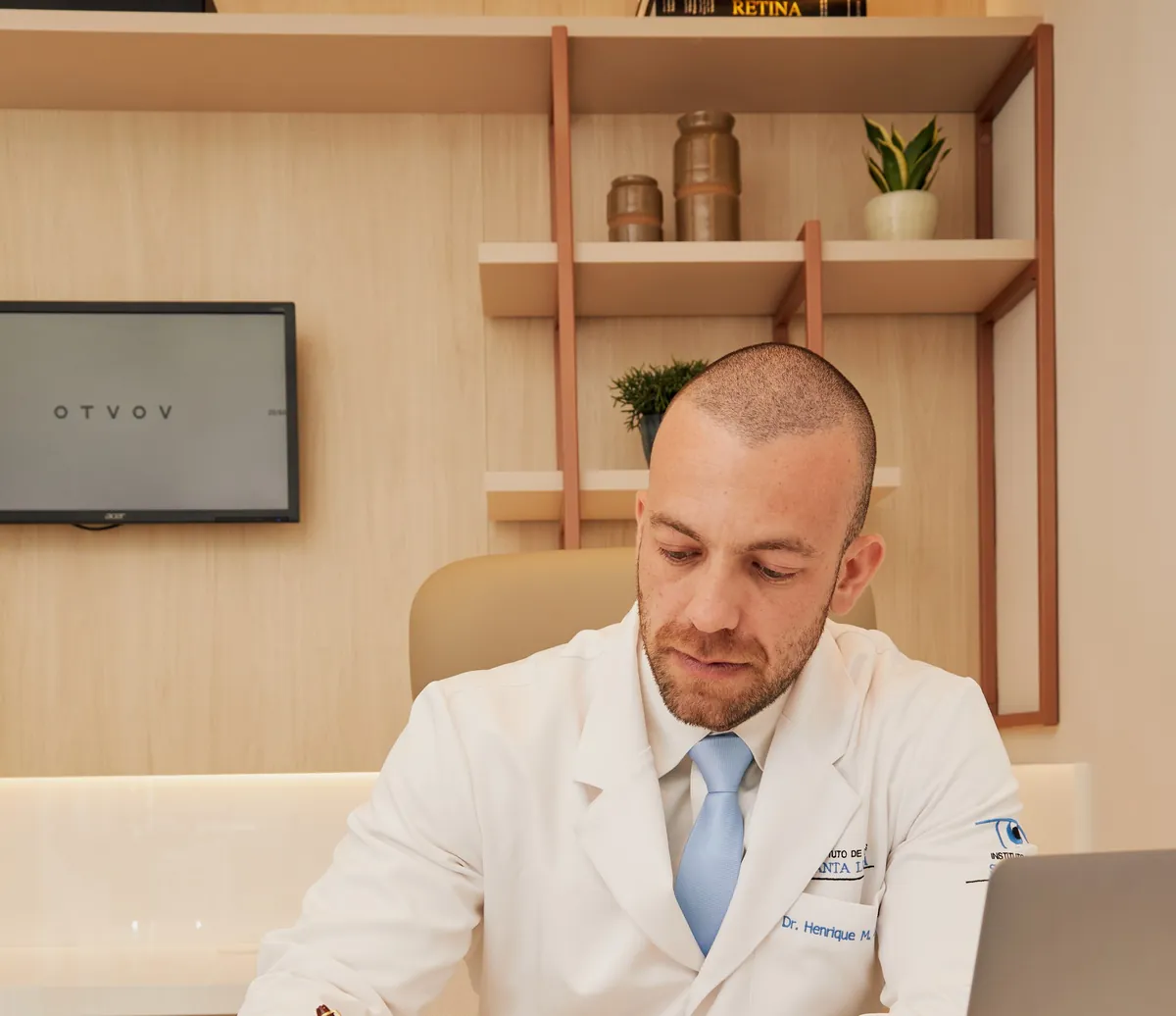 Dr. Henrique Leber, de jaleco, escrevendo em uma mesa de consultório com um notebook ao lado.