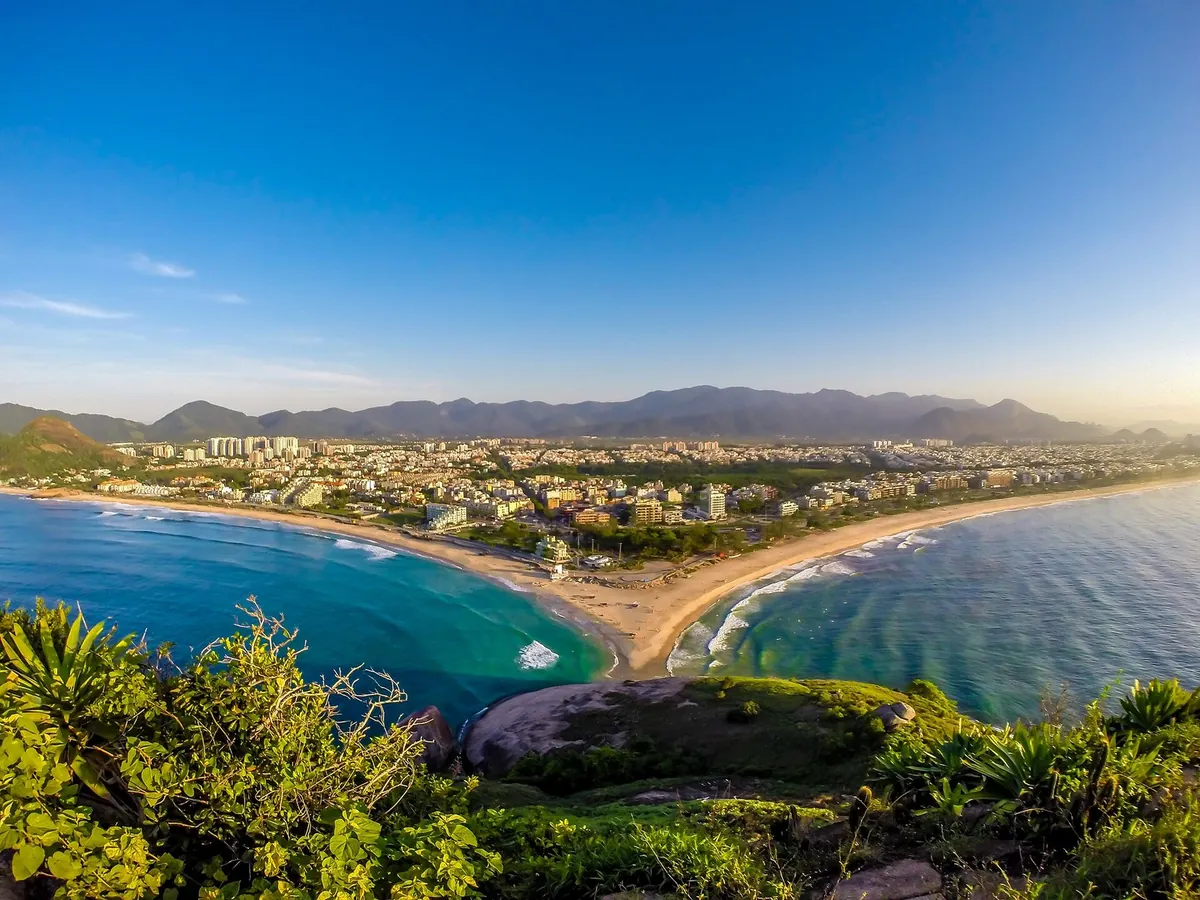 Vista do alto da Praia do Recreio dos Bandeirantes, com a faixa de areia curva, o mar em dois tons de azul e montanhas ao fundo.