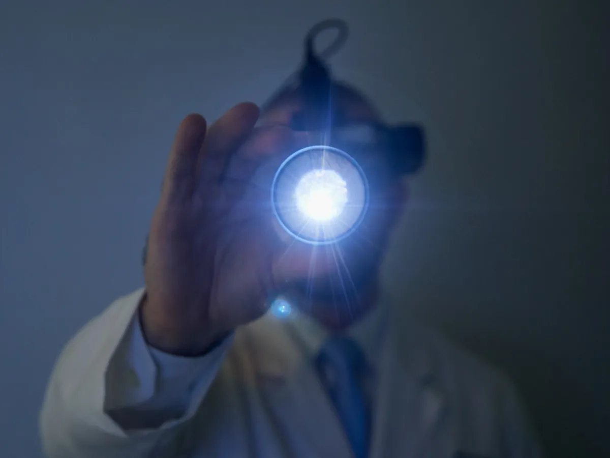 Médico de jaleco segura uma lente diante de uma luz intensa presa à cabeça, equipamento usado para examinar a retina.