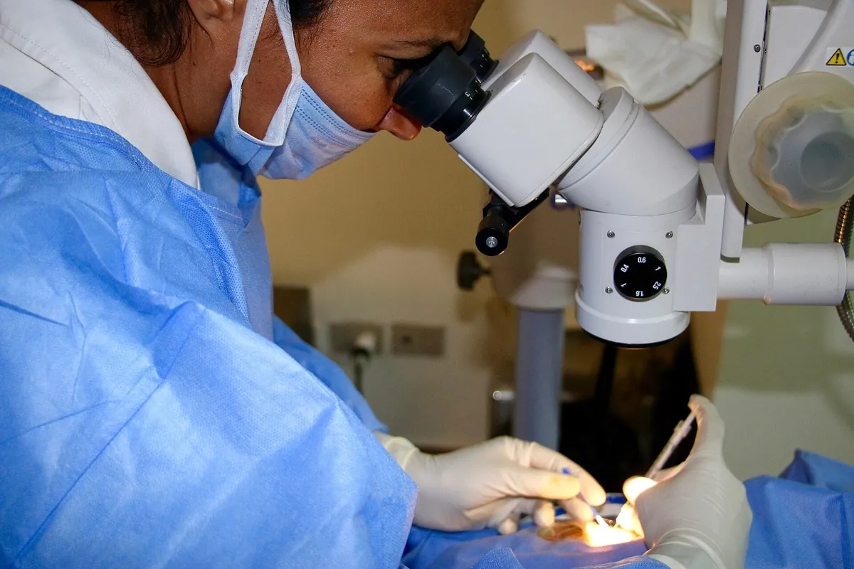 Médica de máscara e avental olhando por um microscópio cirúrgico, enquanto mãos de luvas aplicam uma injeção no olho de uma pessoa.