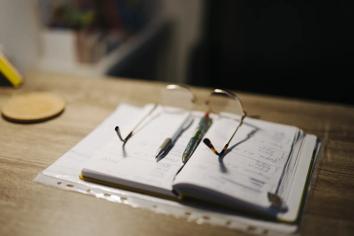 Óculos de leitura abertos e pousados sobre um caderno com canetas em uma mesa.