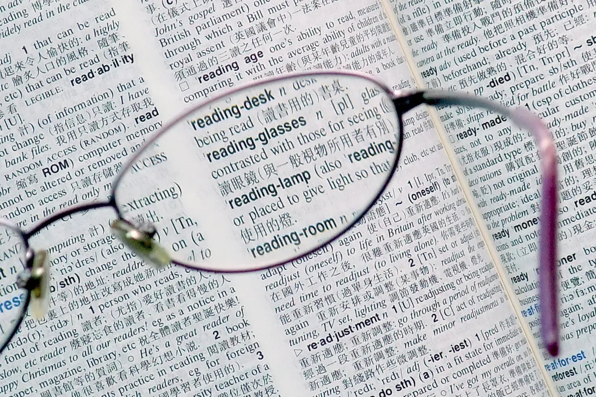 Óculos de leitura apoiados sobre um dicionário aberto, com o verbete 'reading glasses' visível através da lente.