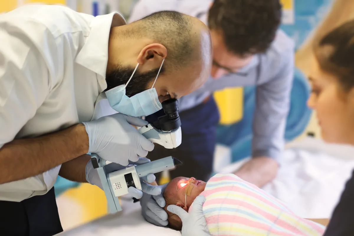 Bebê deitado durante exame ocular, com instrumento portátil dirigido ao olho e mãos enluvadas do examinador.