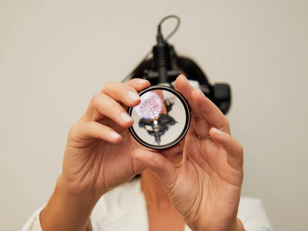 Oftalmologista com uma luz de oftalmoscópio indireto na cabeça, segurando diante do rosto, com as duas mãos, a lente usada no mapeamento de retina.