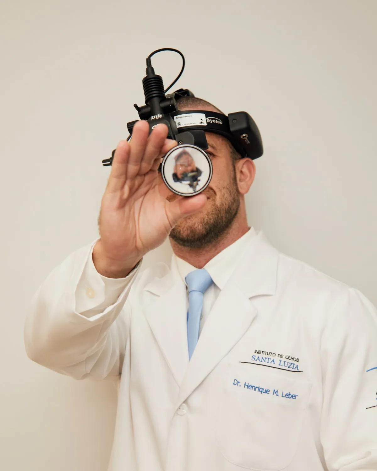 Médico de jaleco segurando uma lente de diagnóstico diante do rosto, com uma luz de cabeça usada em exame de retina.