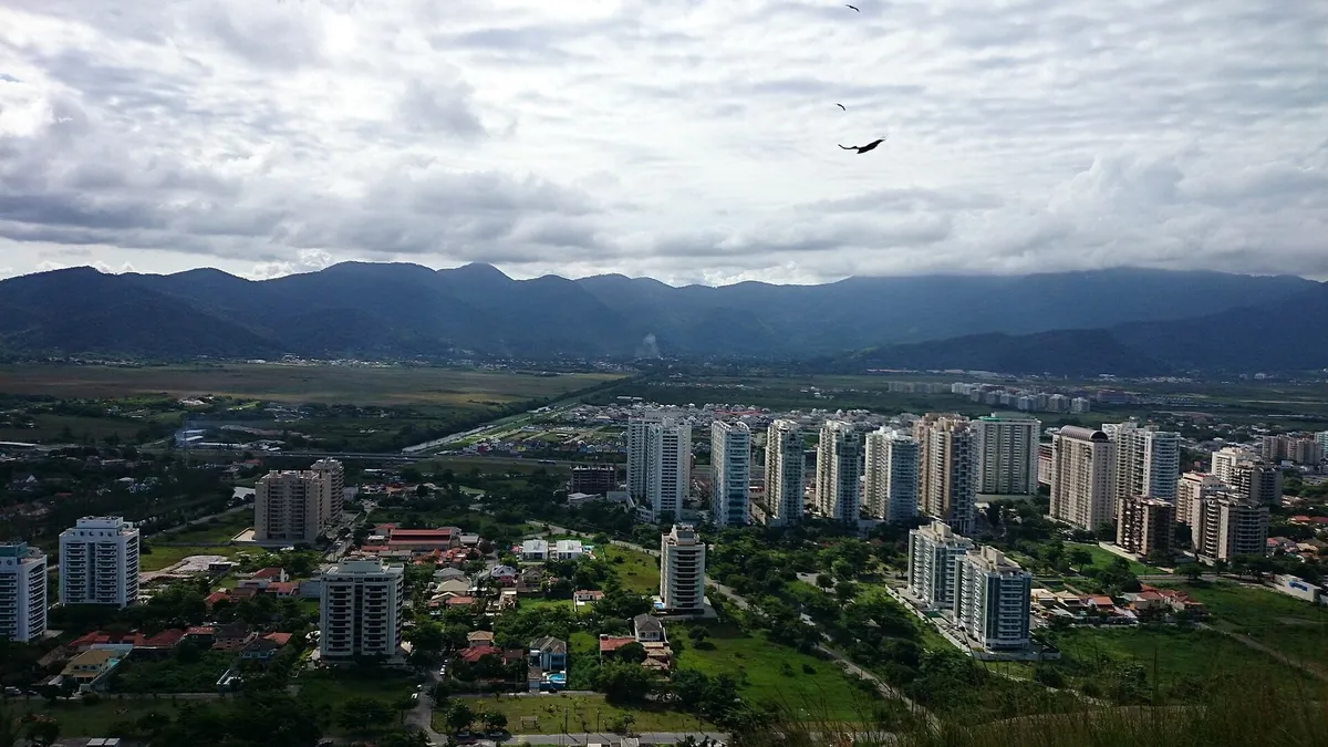 Vista aérea de prédios e ruas arborizadas do Recreio dos Bandeirantes, com montanhas ao fundo e céu nublado.