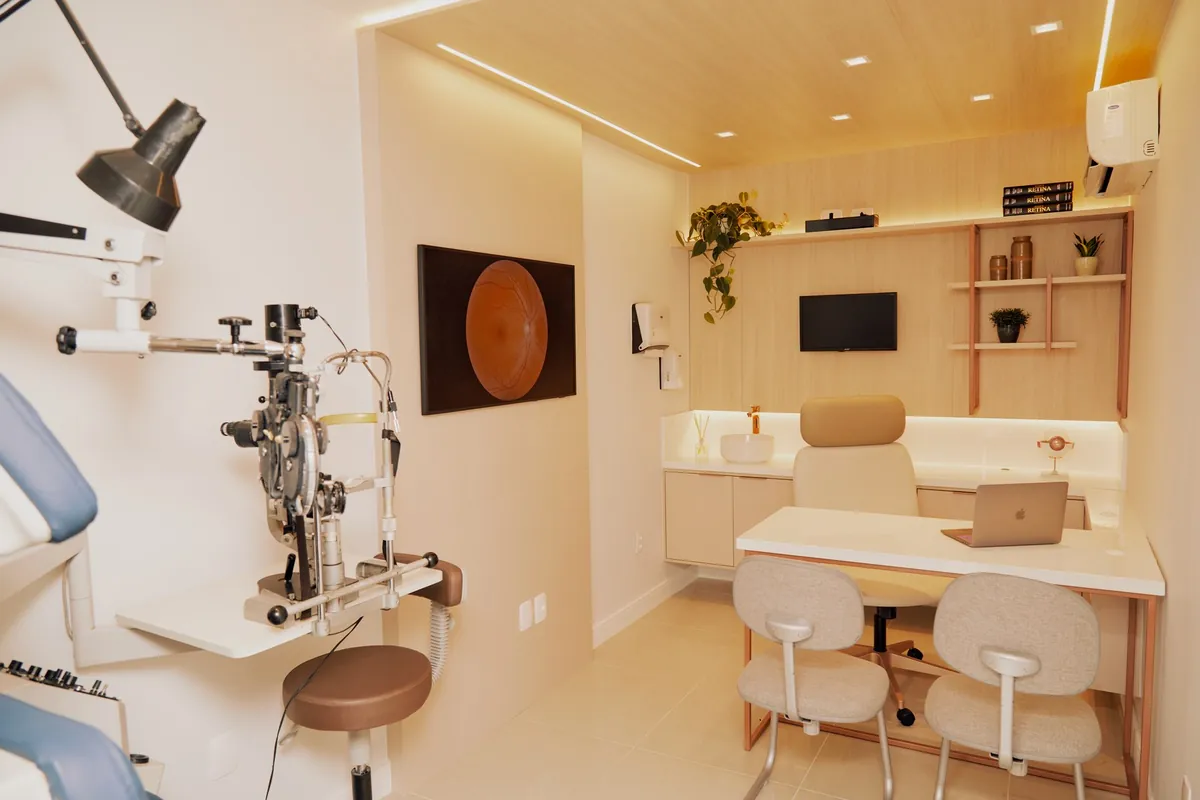 Sala de exame de oftalmologia com lâmpada de fenda, cadeira de exame, mesa de consulta e um quadro de retina na parede.