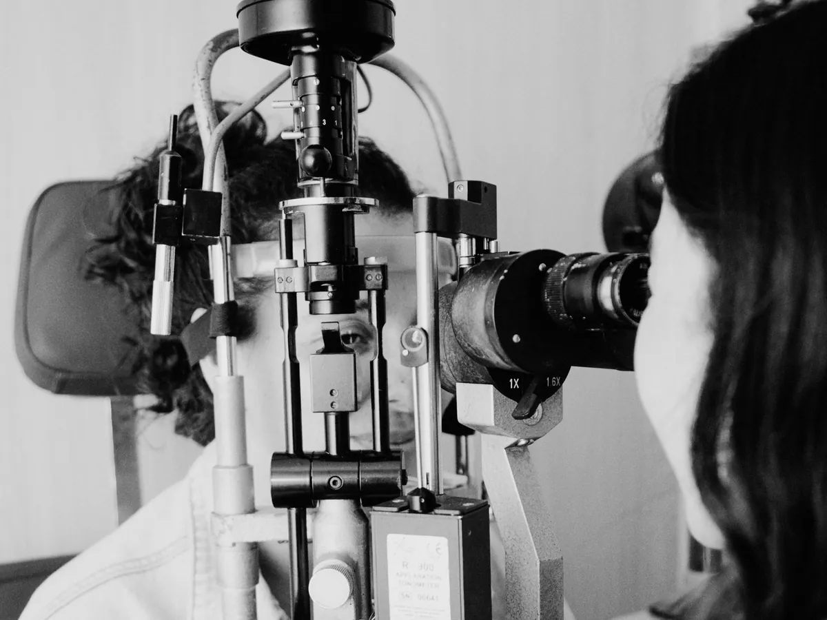 Fotografia em preto e branco de uma médica examinando o olho de uma pessoa em uma lâmpada de fenda.