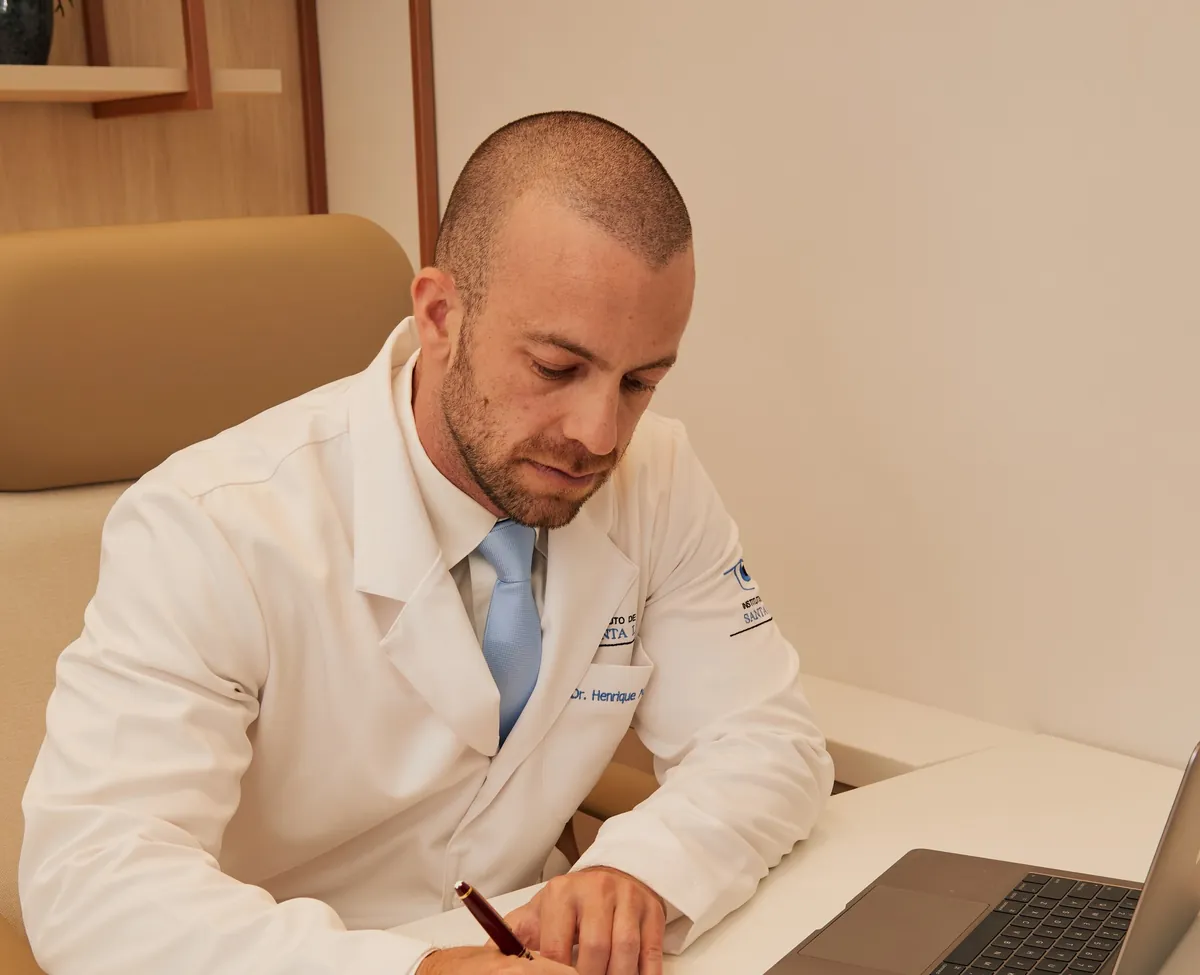 Dr. Henrique Leber, de jaleco, escrevendo em uma mesa de consultório com um notebook ao lado.