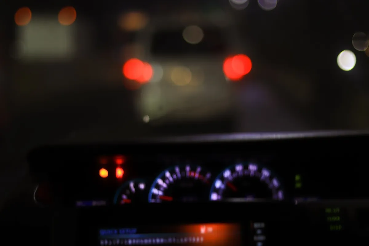 Painel de um carro e luzes vermelhas e brancas de outros veículos borradas ao fundo, em uma estrada à noite.
