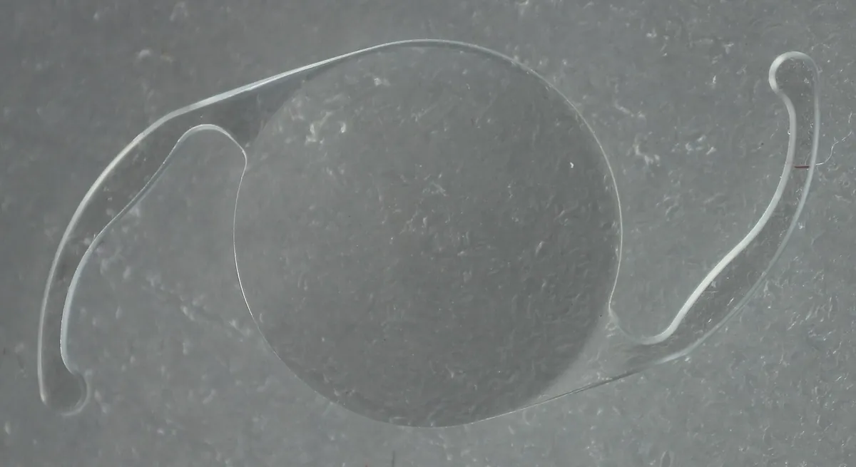 Lente intraocular transparente, com duas hastes finas, fotografada de perto sobre fundo cinza.