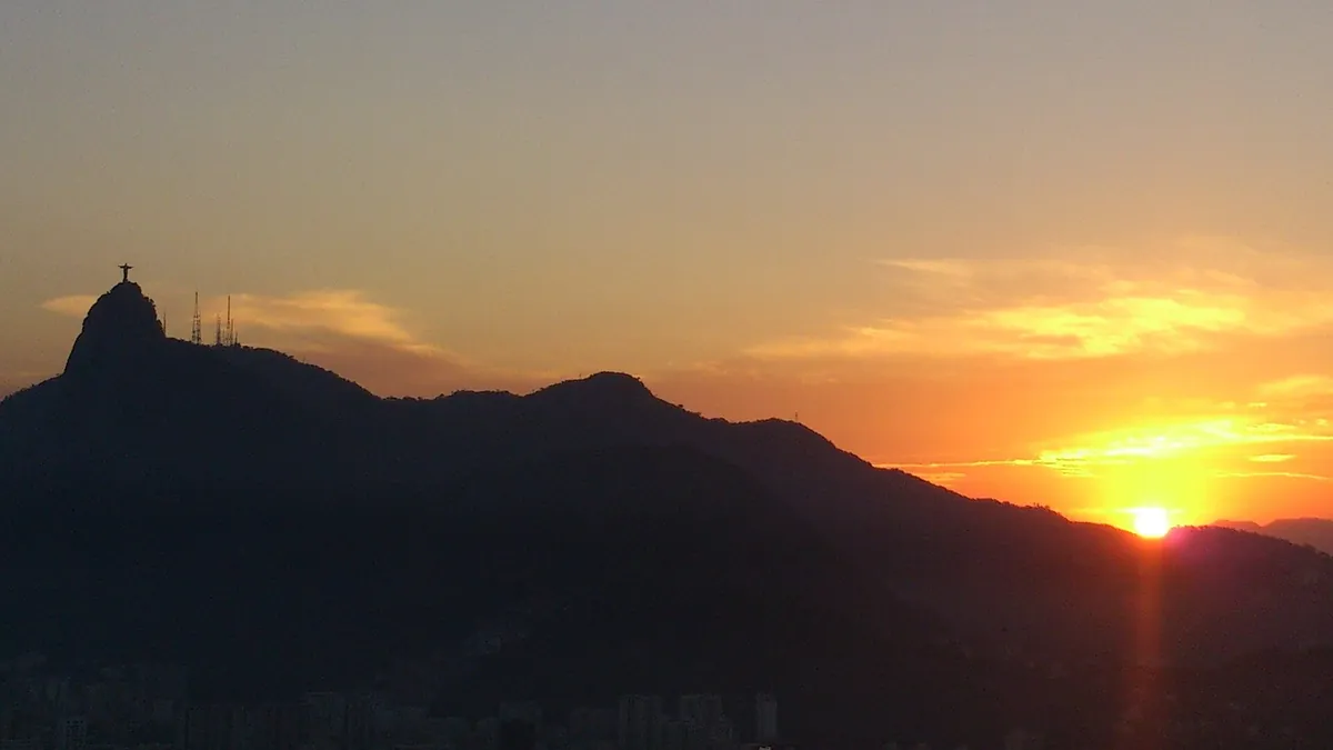 Pôr do sol alaranjado atrás das montanhas do Rio de Janeiro, com o Cristo Redentor à esquerda.