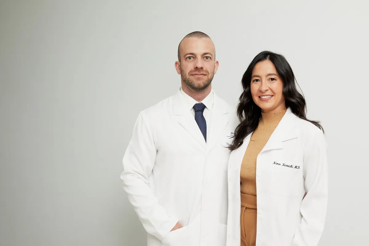 Dr. Henrique Leber e Dra. Nina Konichi, de jaleco branco, lado a lado diante de um fundo claro.